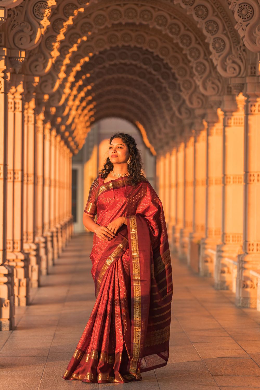 A person wearing a red silk sari poses elegantly in an ornate columned corridor lit by warm golden light.