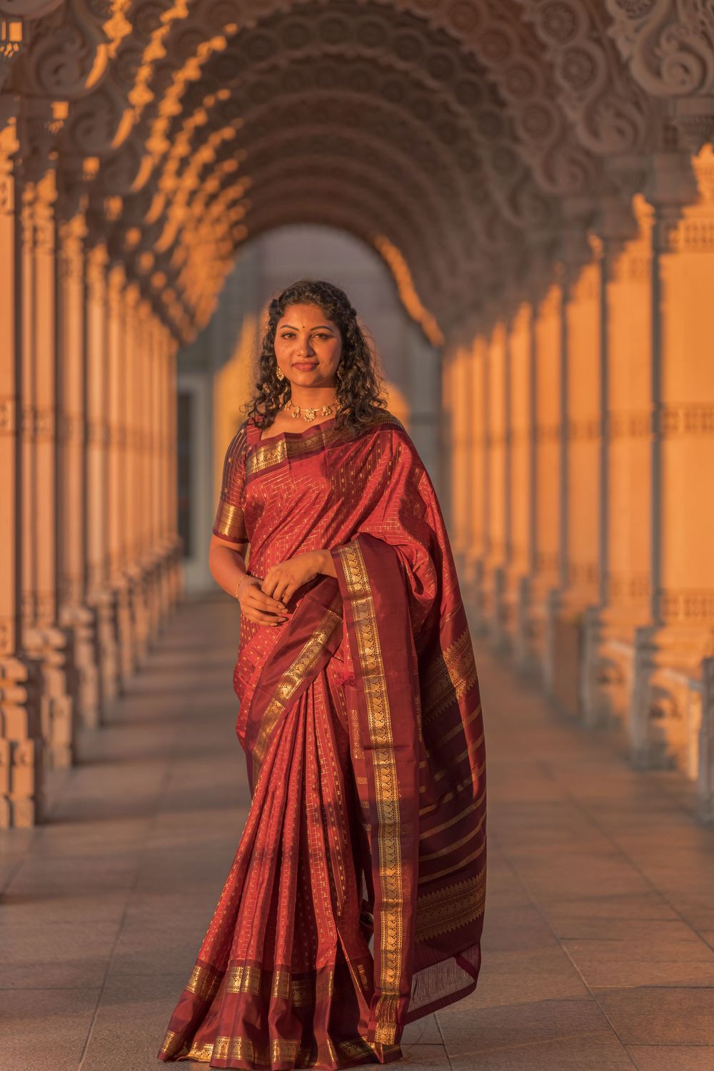 A person in a red sari poses elegantly in a grand colonnade corridor with warm sunset lighting.