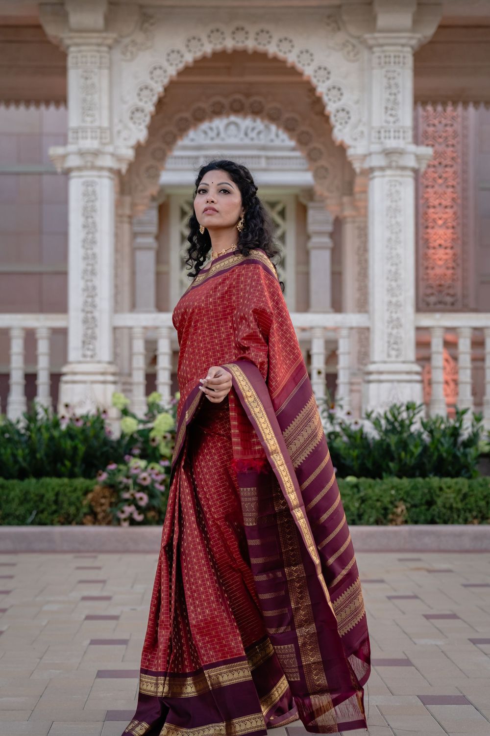 Woman in traditional red and maroon Indian attire poses gracefully against ornate architectural backdrop.