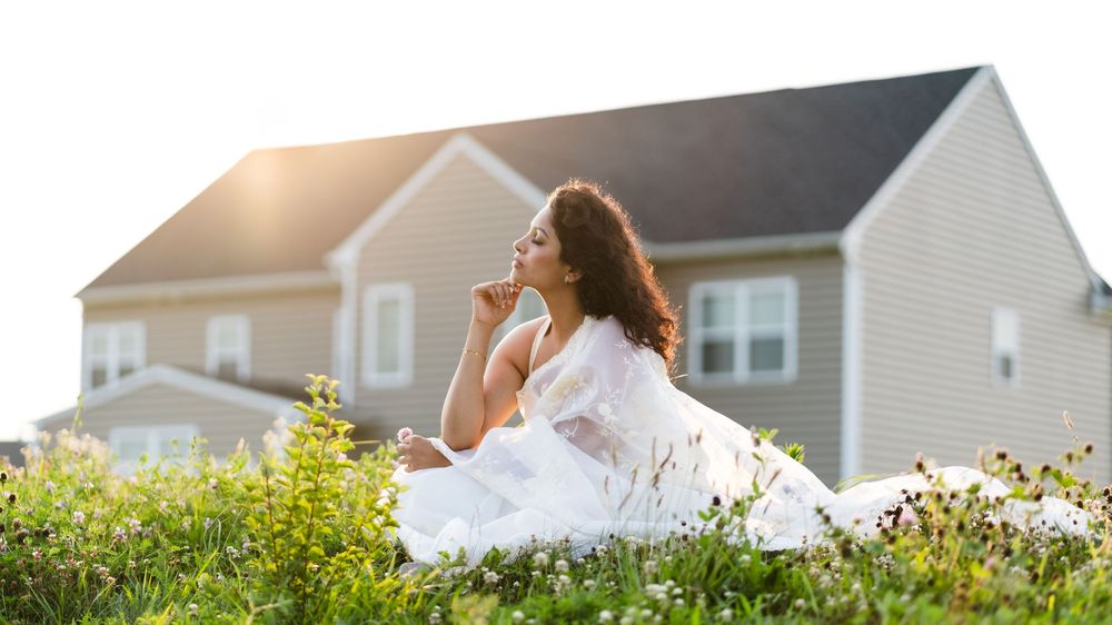 A person in a white dress sits thoughtfully in a sunny meadow with wildflowers, a suburban house in the background.
