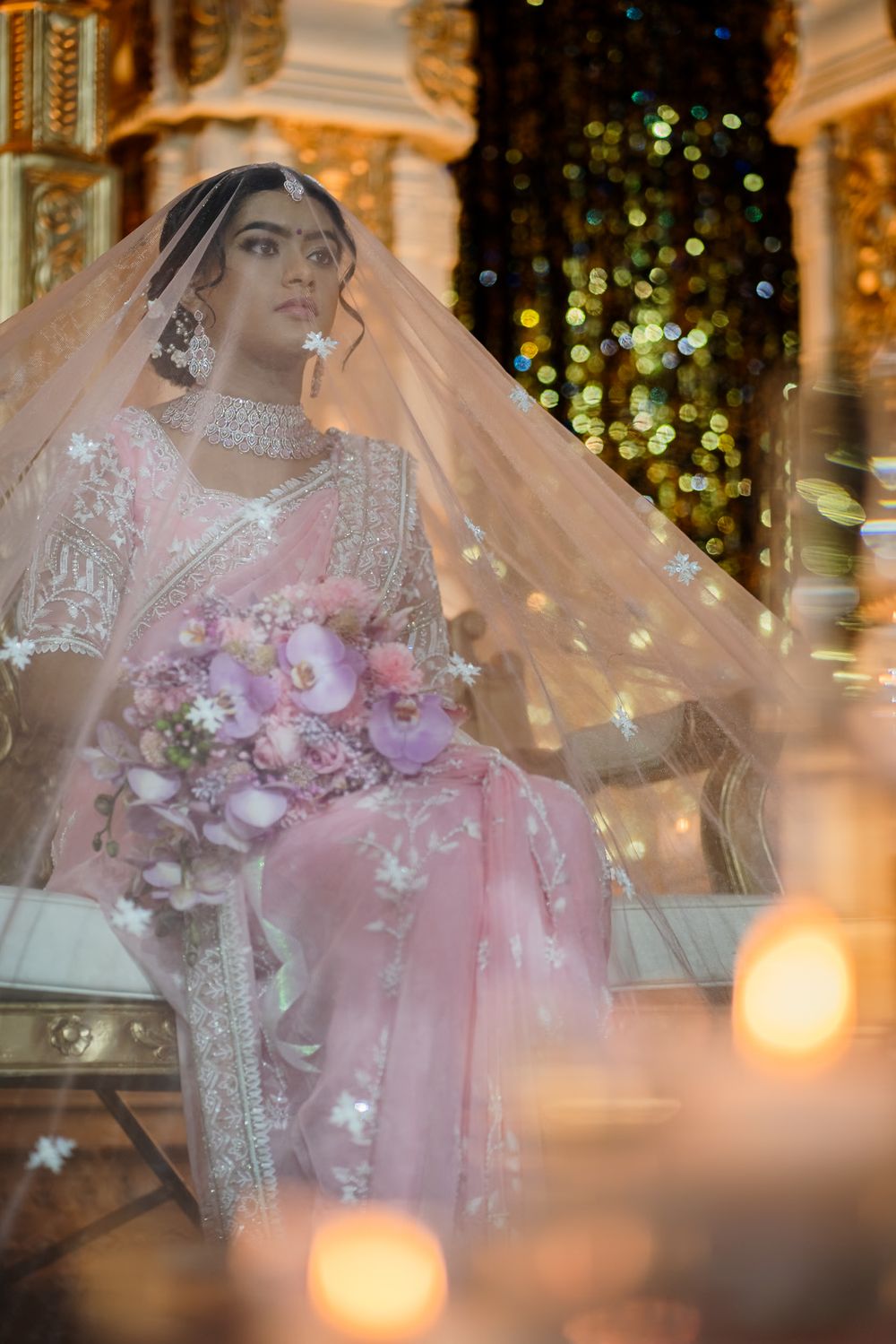 A bride in a pink and white traditional South Asian wedding attire holds a pink floral bouquet against a bokeh background.