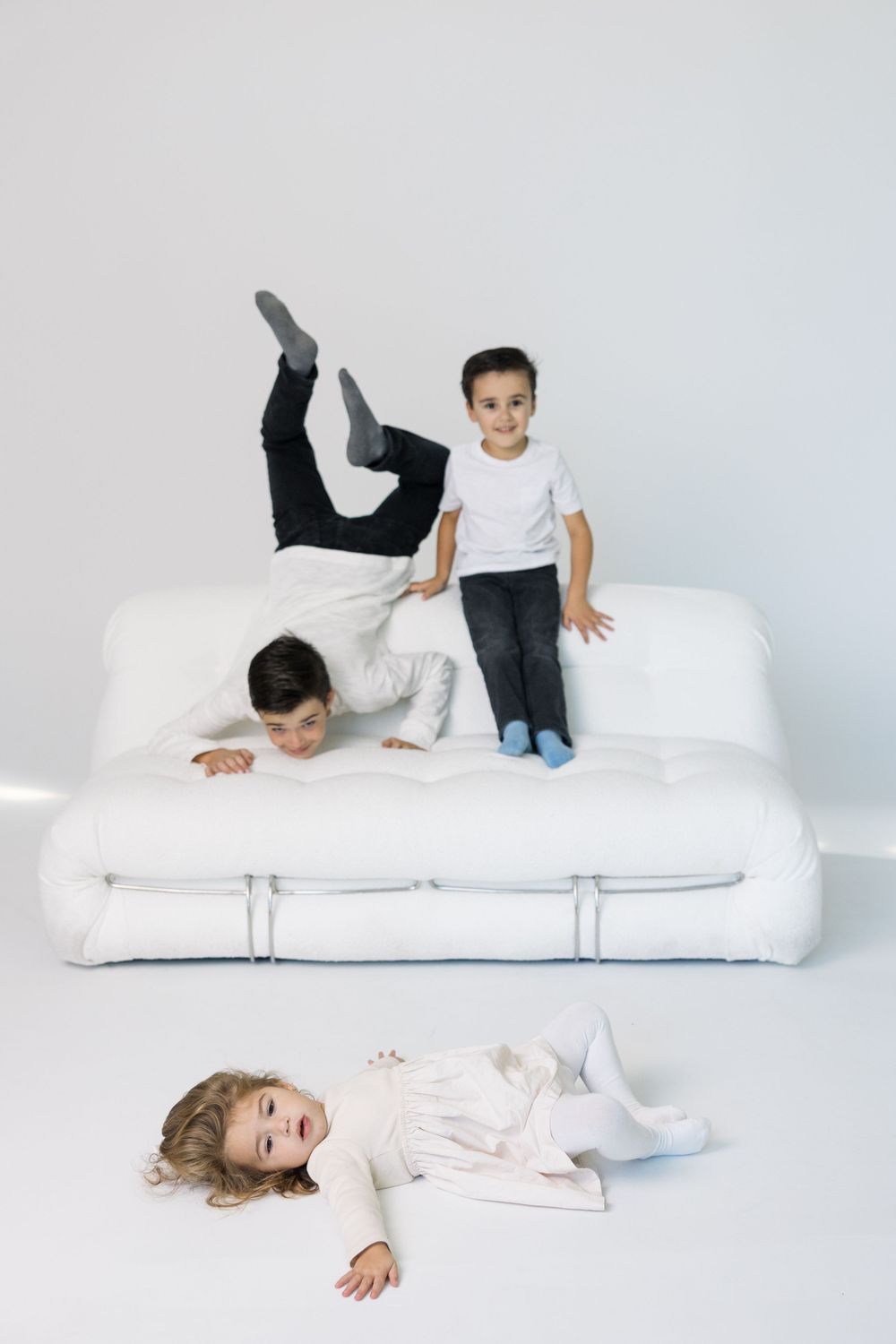 Children playing and posing playfully on a white couch in a minimalist setting.