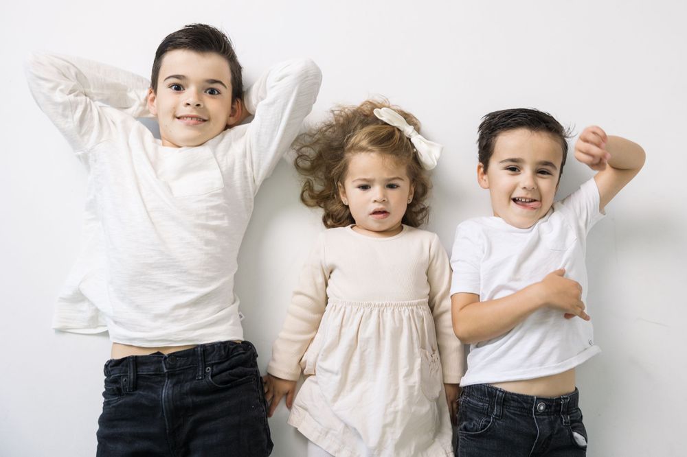 Three young children in white clothing pose against a light background.