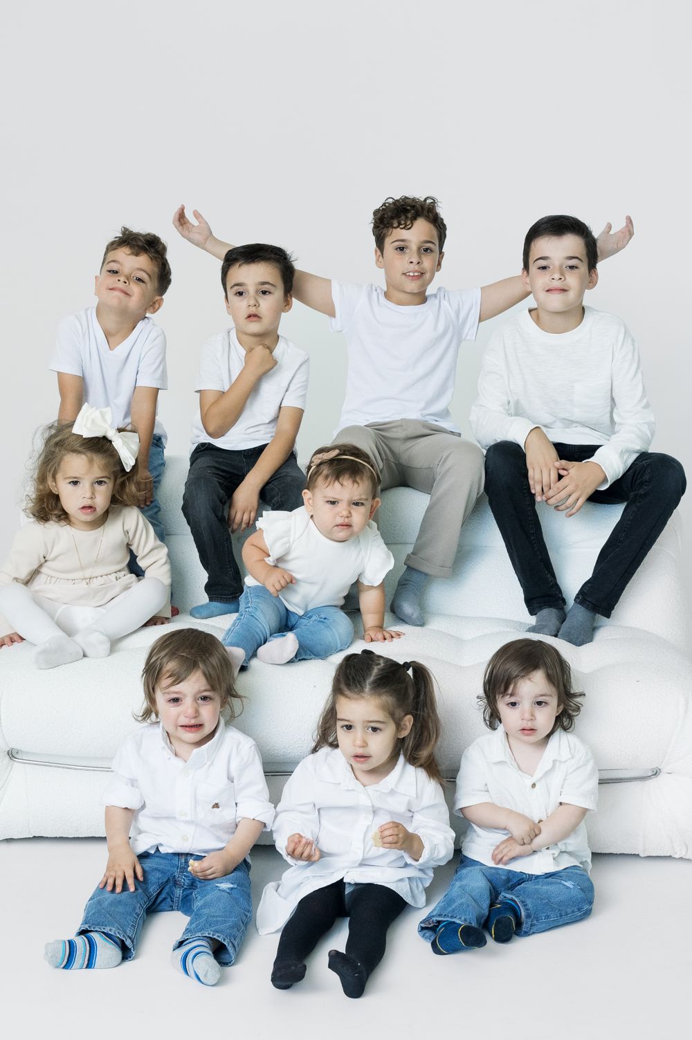 Nine children arranged in two rows on white cushions wearing casual clothing.