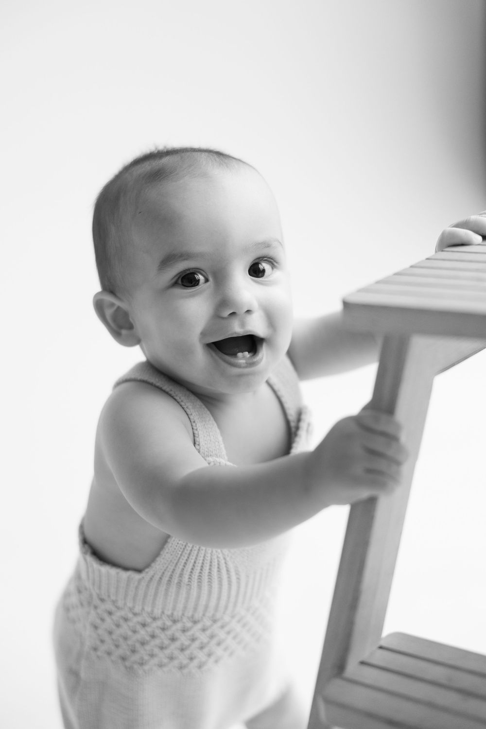 A happy baby in white clothing holds onto furniture while standing, shown in a series of black and white photos.