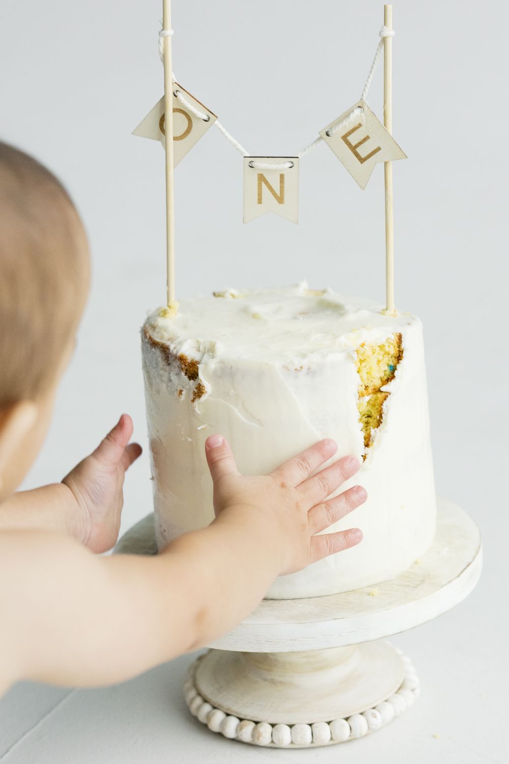 Small hands reach for a white frosted cake on a pedestal stand with gold letter decorations spelling ONE on top.