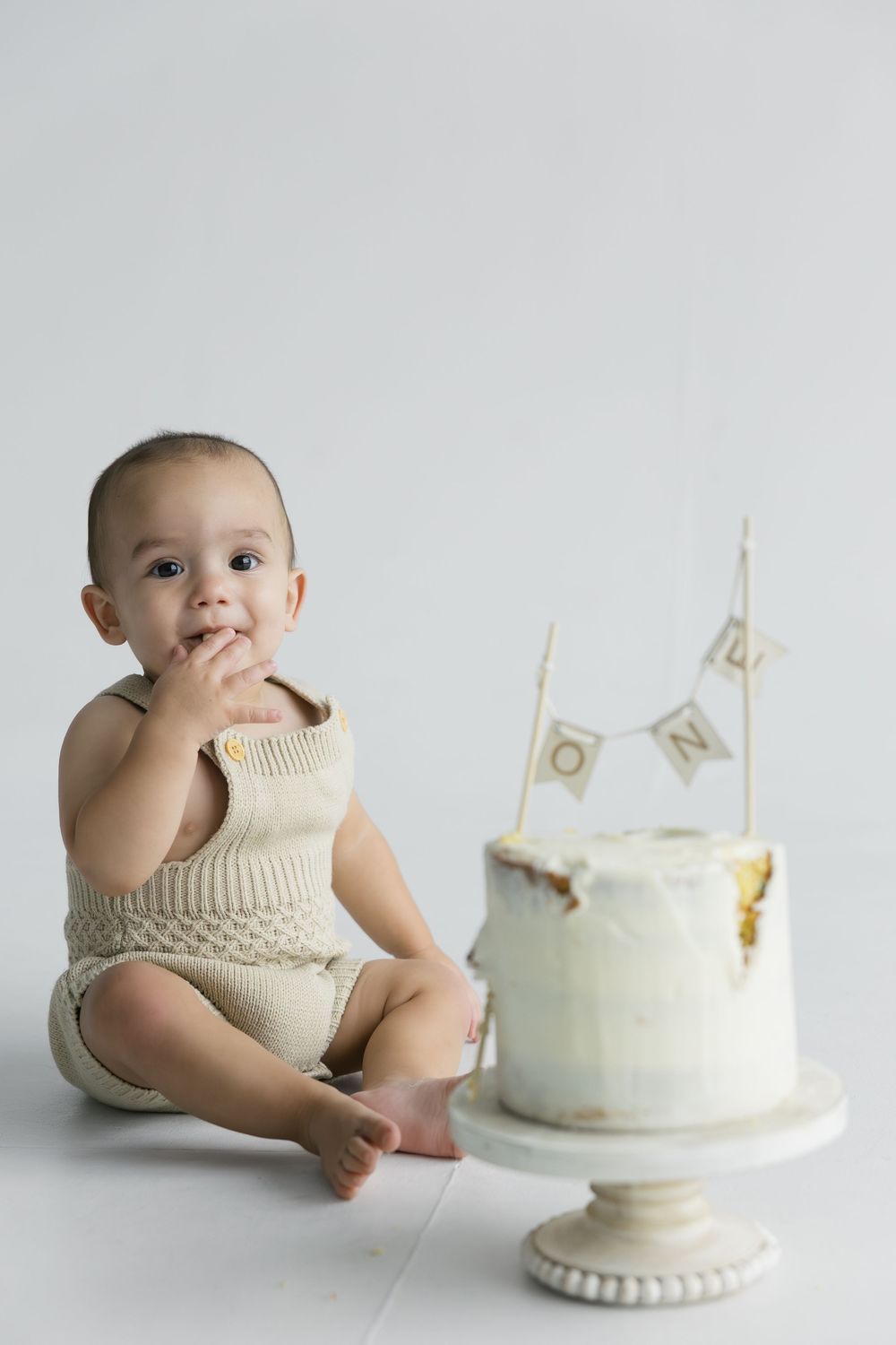 Minimalist photo series showing a baby in cream-colored outfit beside an elegant white birthday cake on a pedestal.