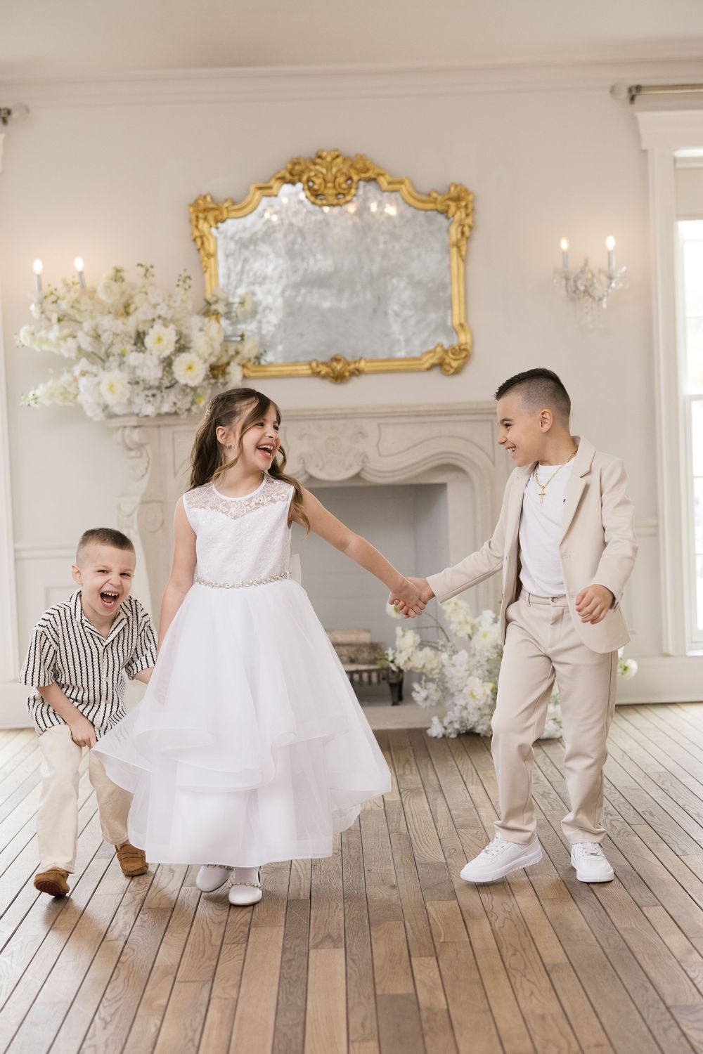 Three children in formal wear dance joyfully in an elegant white room with a gold mirror and fireplace.