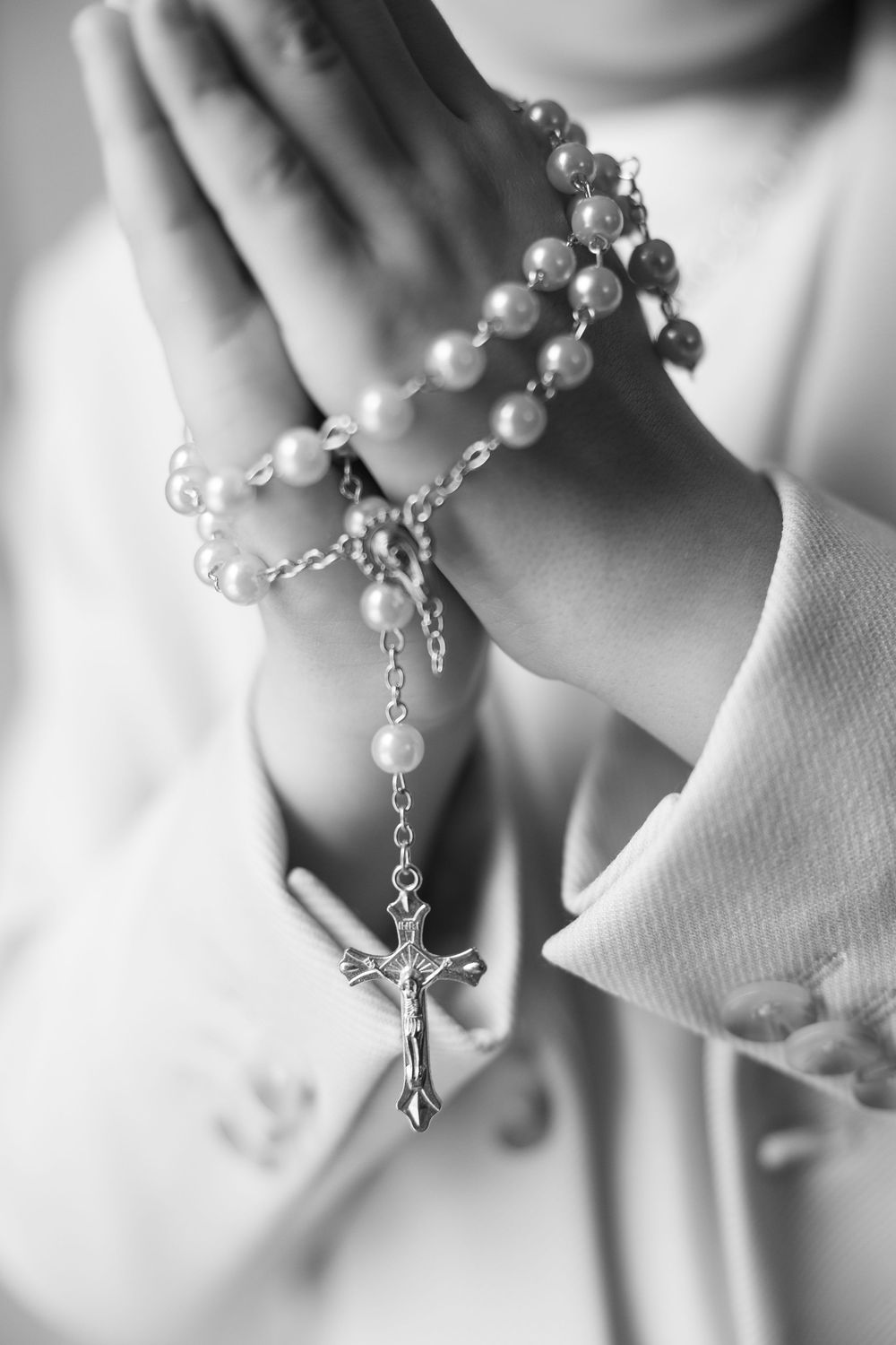Close-up black and white photo of child hands clasped in prayer holding a pearl rosary with crucifix.