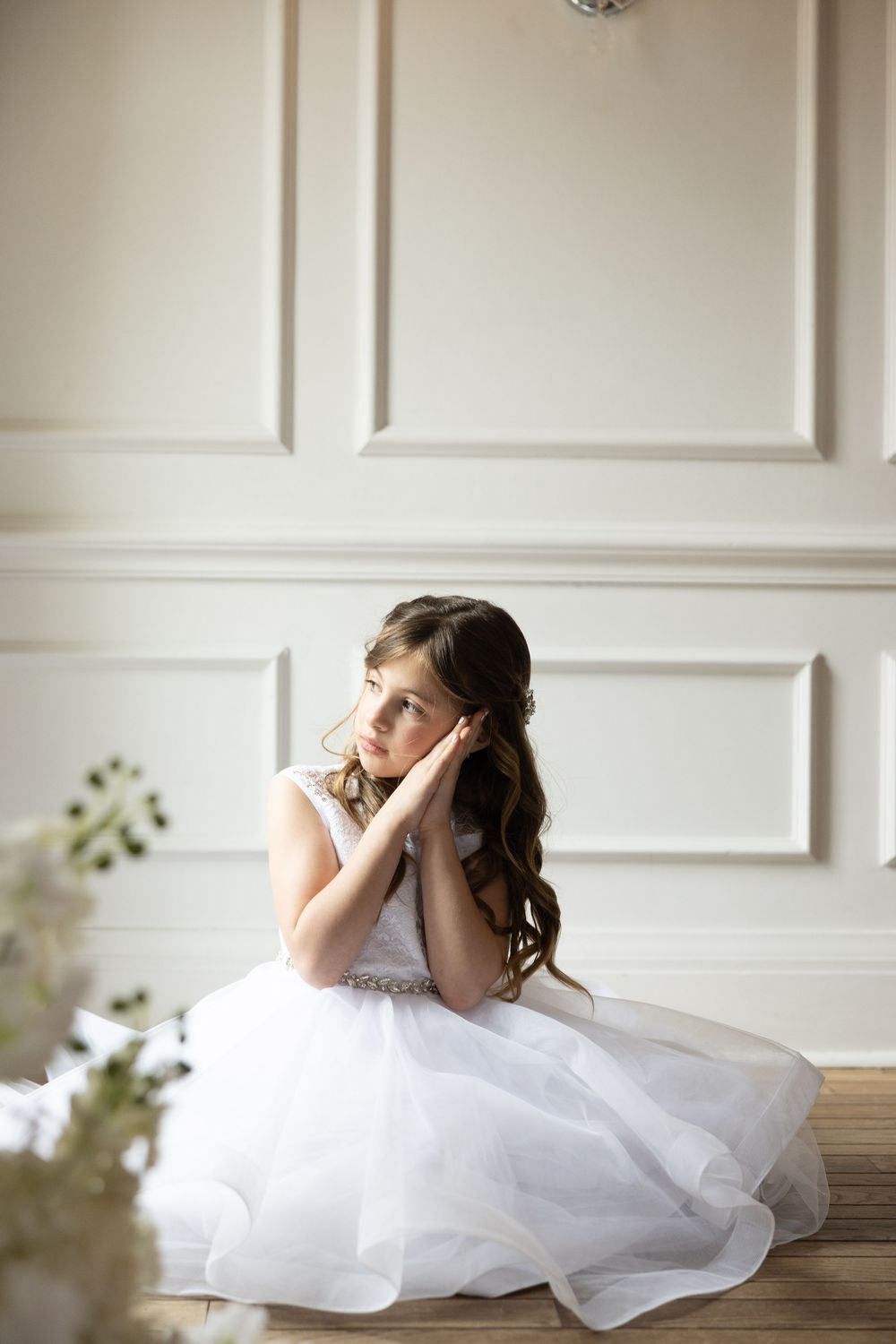 Young girl in white tulle dress sits gracefully on floor, hands clasped near face, in elegant white paneled room.