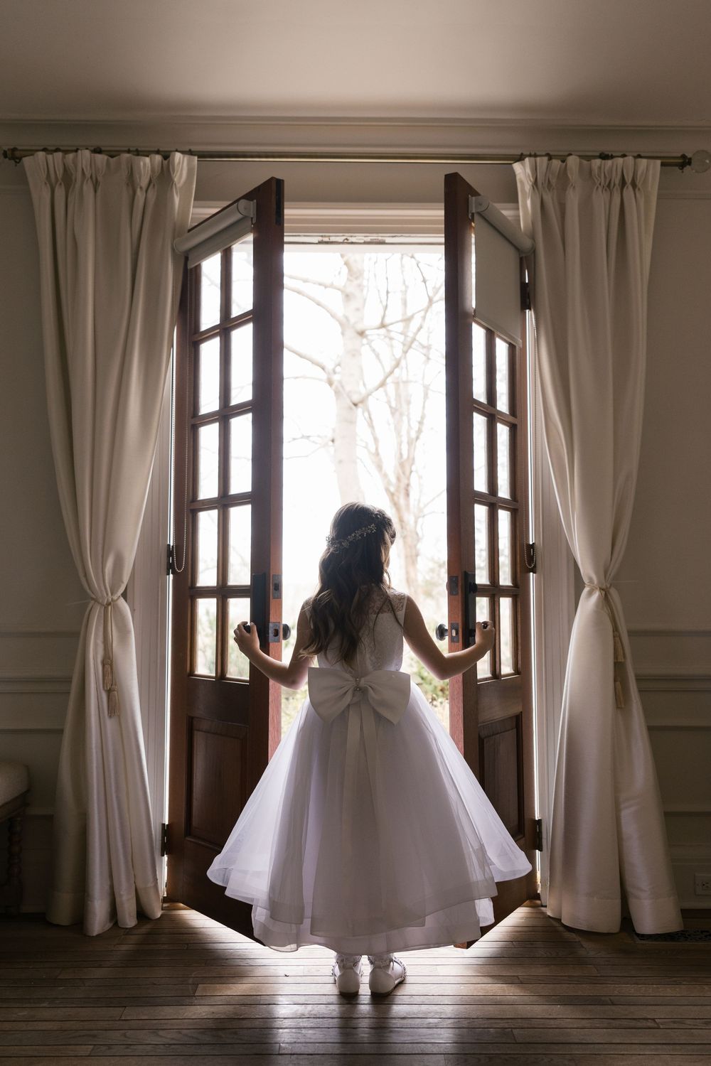 Young girl in white dress with bow stands at open French doors, viewed from behind, with white curtains and natural light.