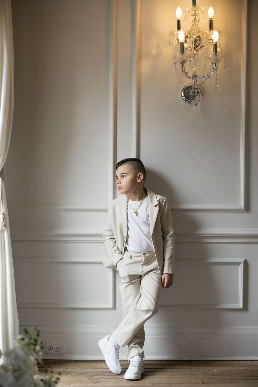A young boy in a beige suit and white sneakers leans against an elegant white wall beneath a crystal chandelier.