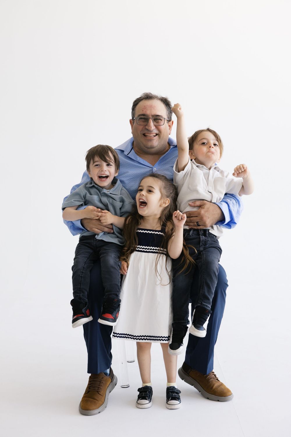 A smiling father sits holding two young boys while a girl stands between his knees, all posing happily against a white background.