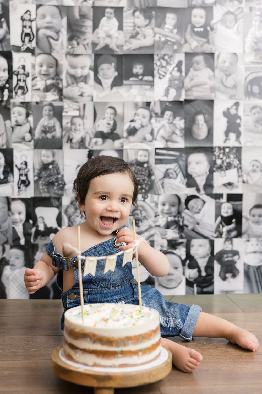 Smiling baby in denim overalls celebrating first birthday with cake, surrounded by black and white photo collage backdrop.