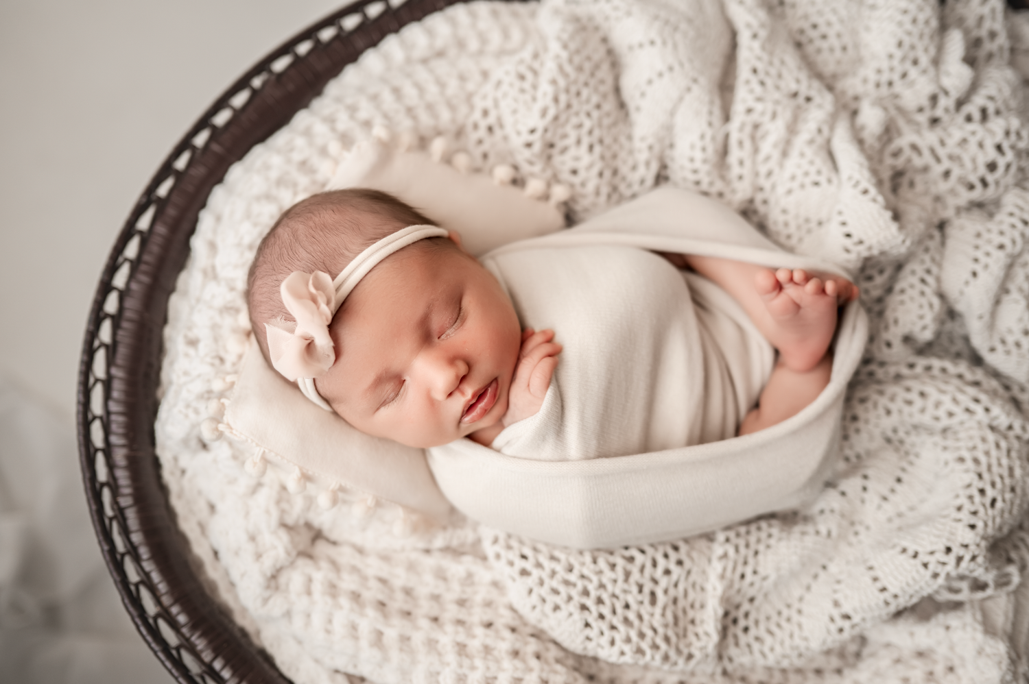 Baby in knit hat and gown lying on bed while parents kiss in the background.