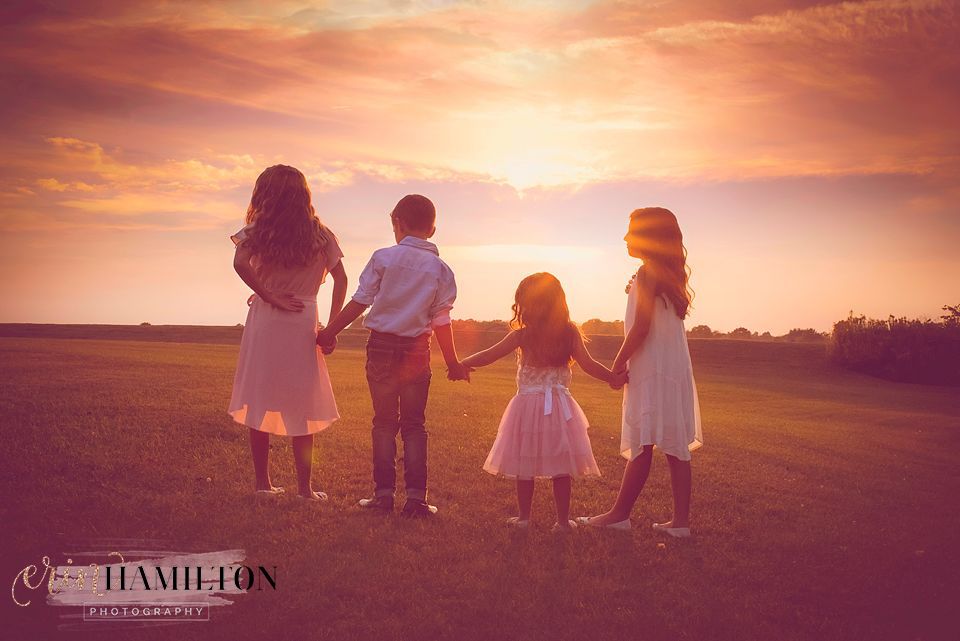 Four silhouettes hold hands while watching a vibrant sunset over an open field.