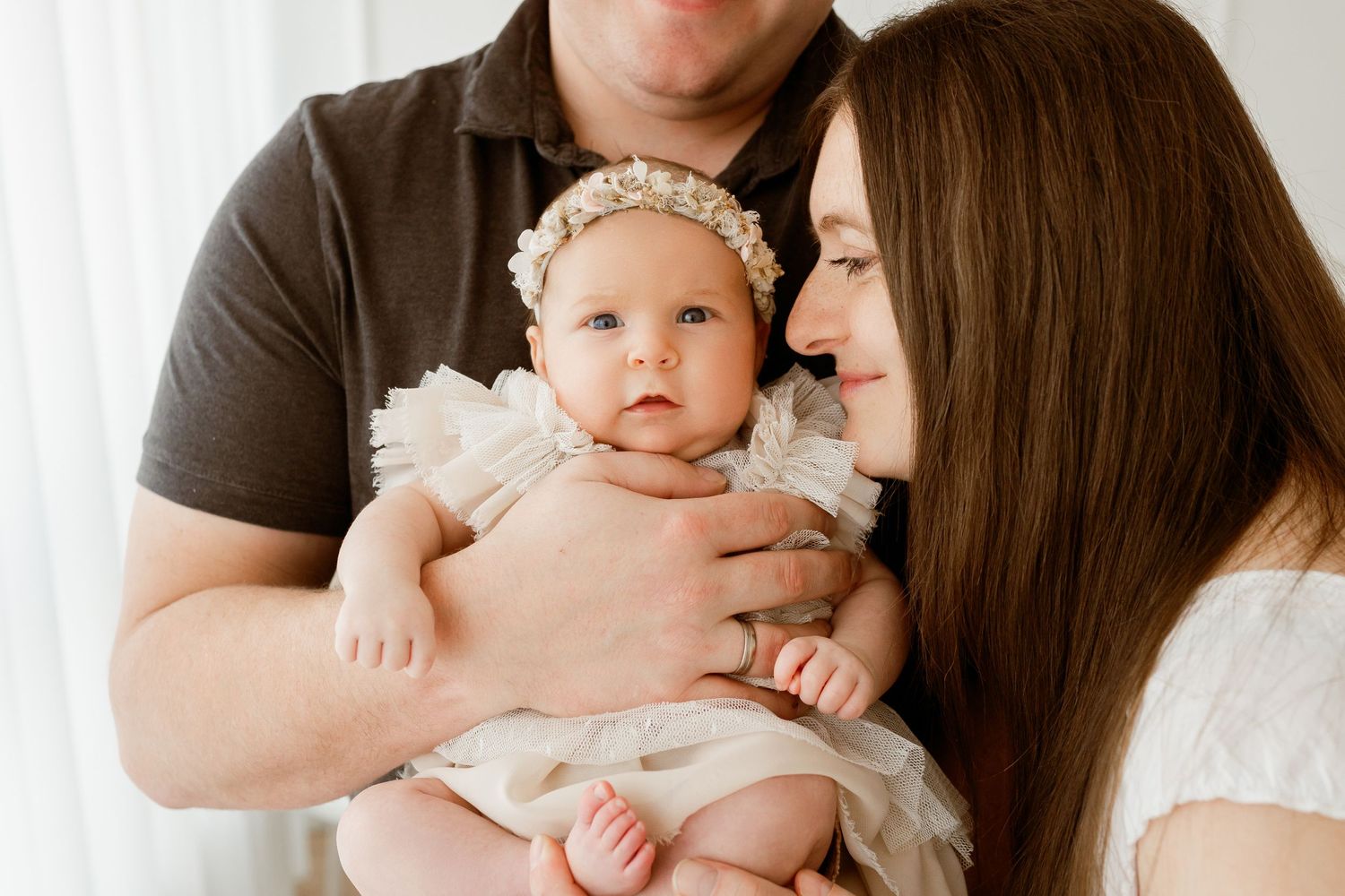 Baby in white dress being held and cuddled by parents against neutral background.