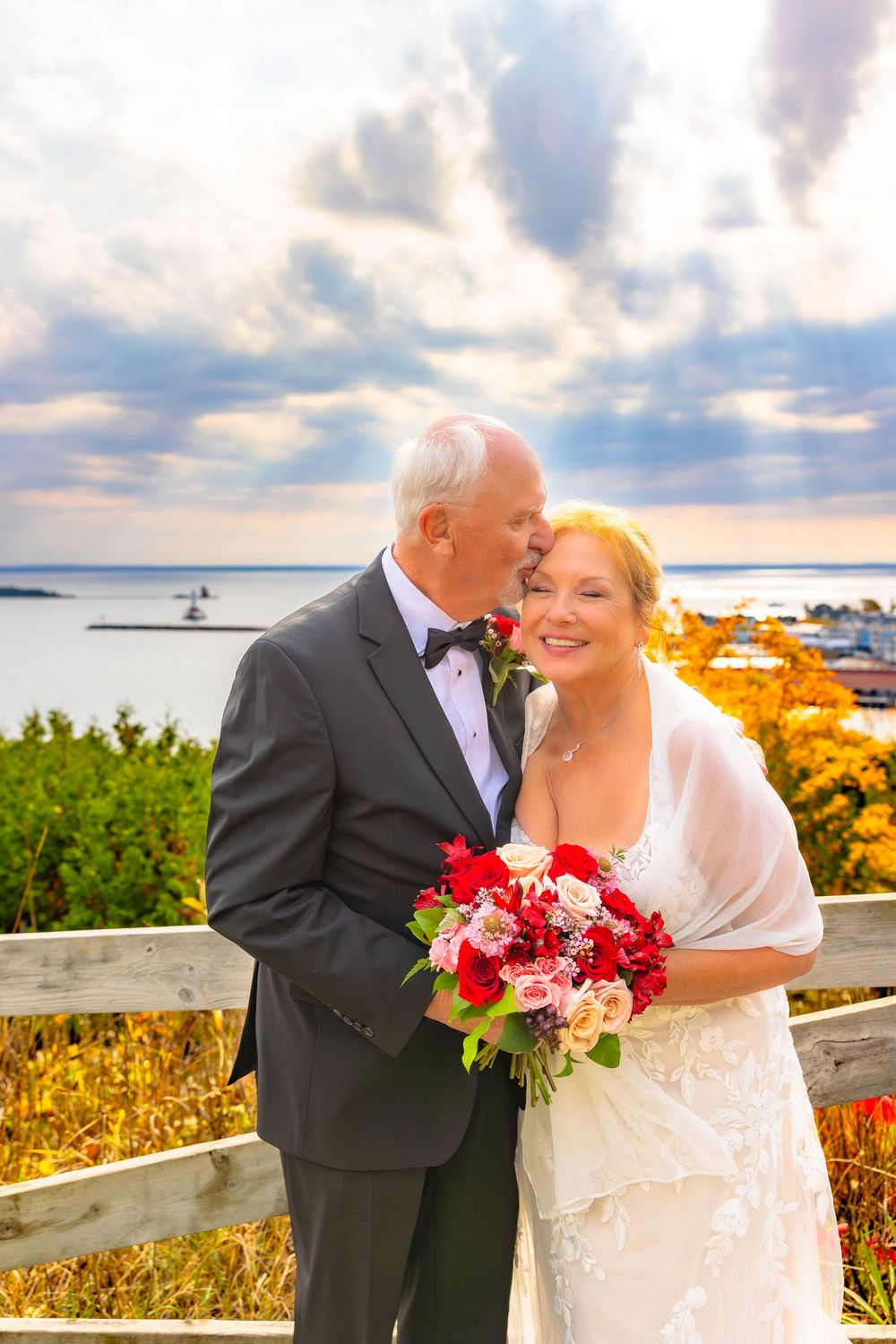 Couple sharing romantic moment by the ocean with red and pink bouquet during sunset ceremony.