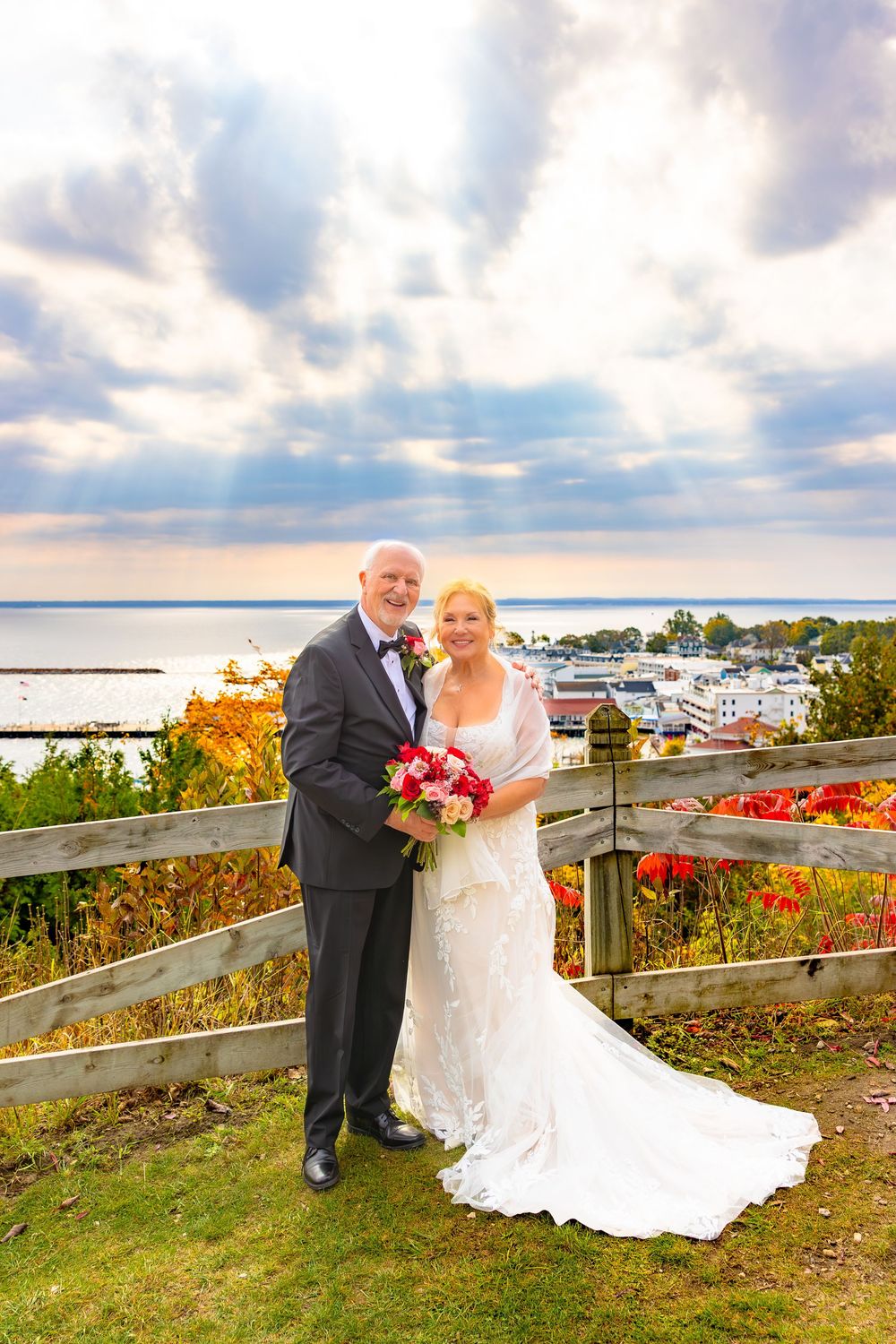 A couple poses together on a scenic overlook deck with fall foliage and boats in the harbor during a sunset wedding.
