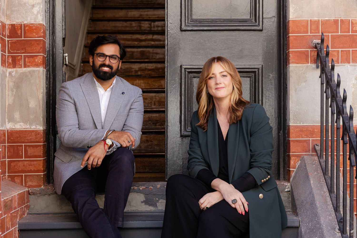 Two business professionals sit casually on gray stone steps in front of a brick building entrance.