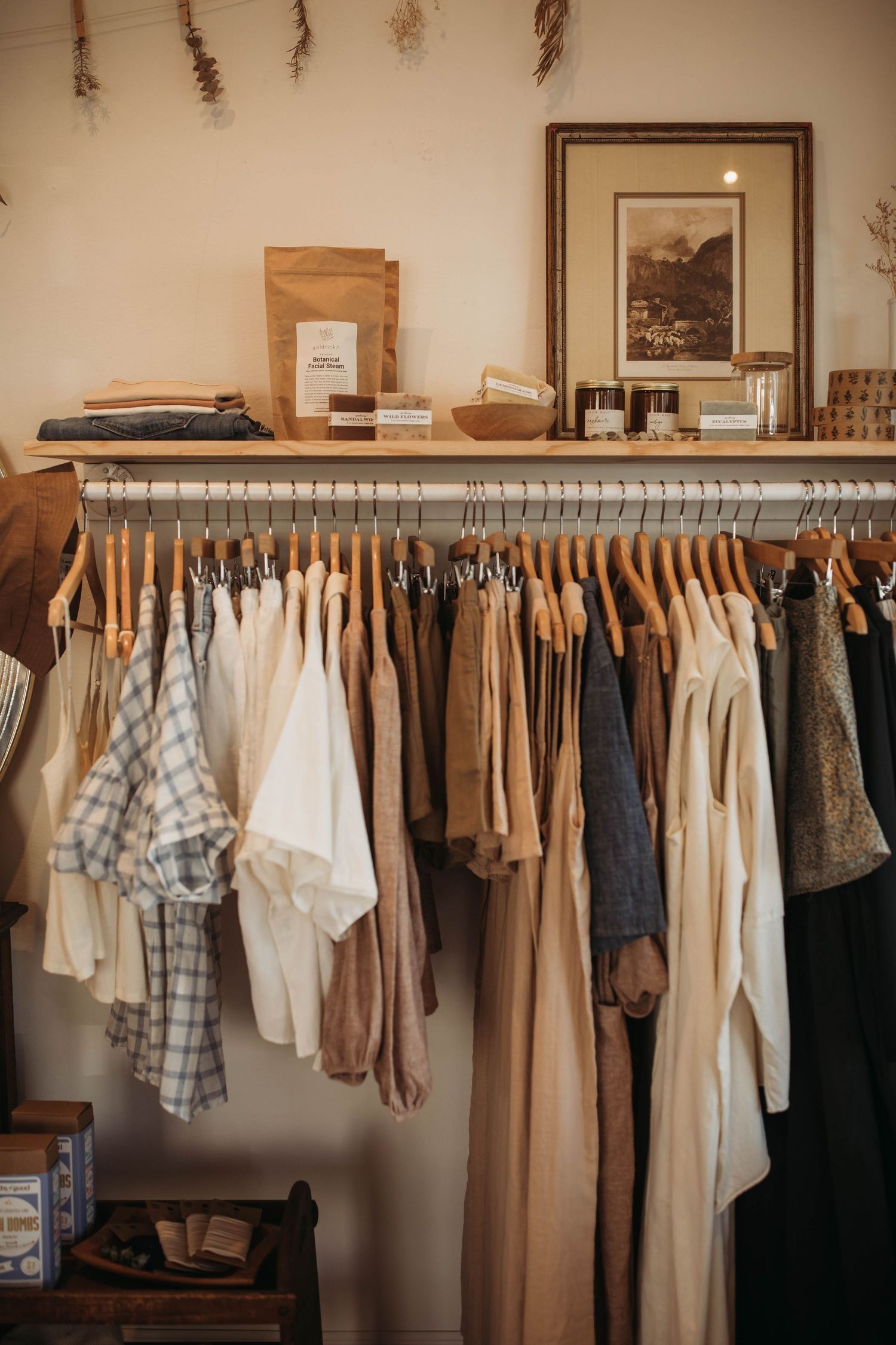 A minimalist clothing rack displays earth-toned garments and neutral colors with rustic decor on shelf above.