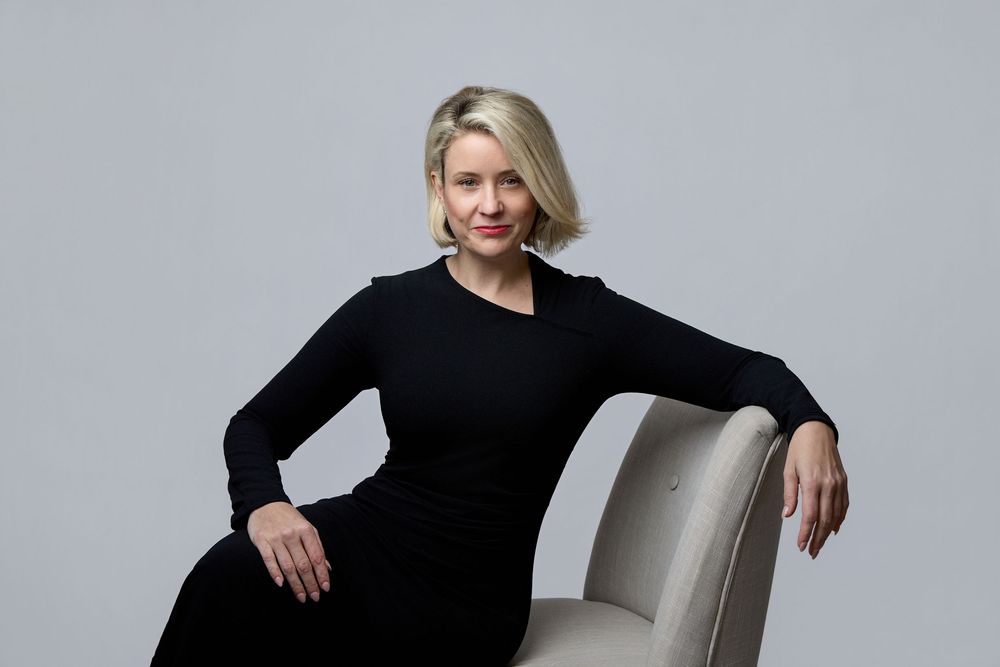 Woman in black dress poses elegantly on white chair against gray backdrop in professional photo series.