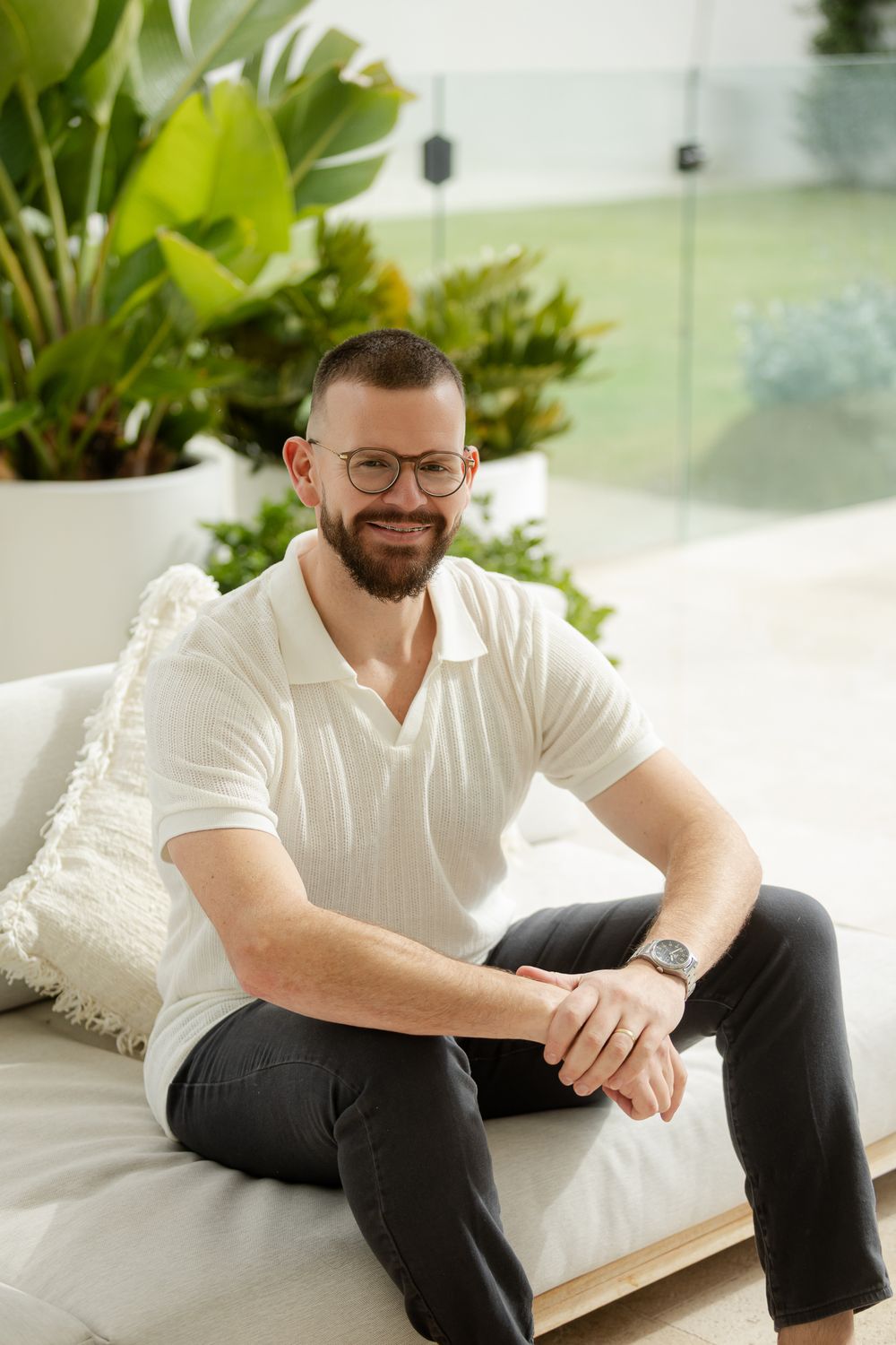 A person in glasses and a white polo shirt sits casually on a white couch with indoor plants in the background.
