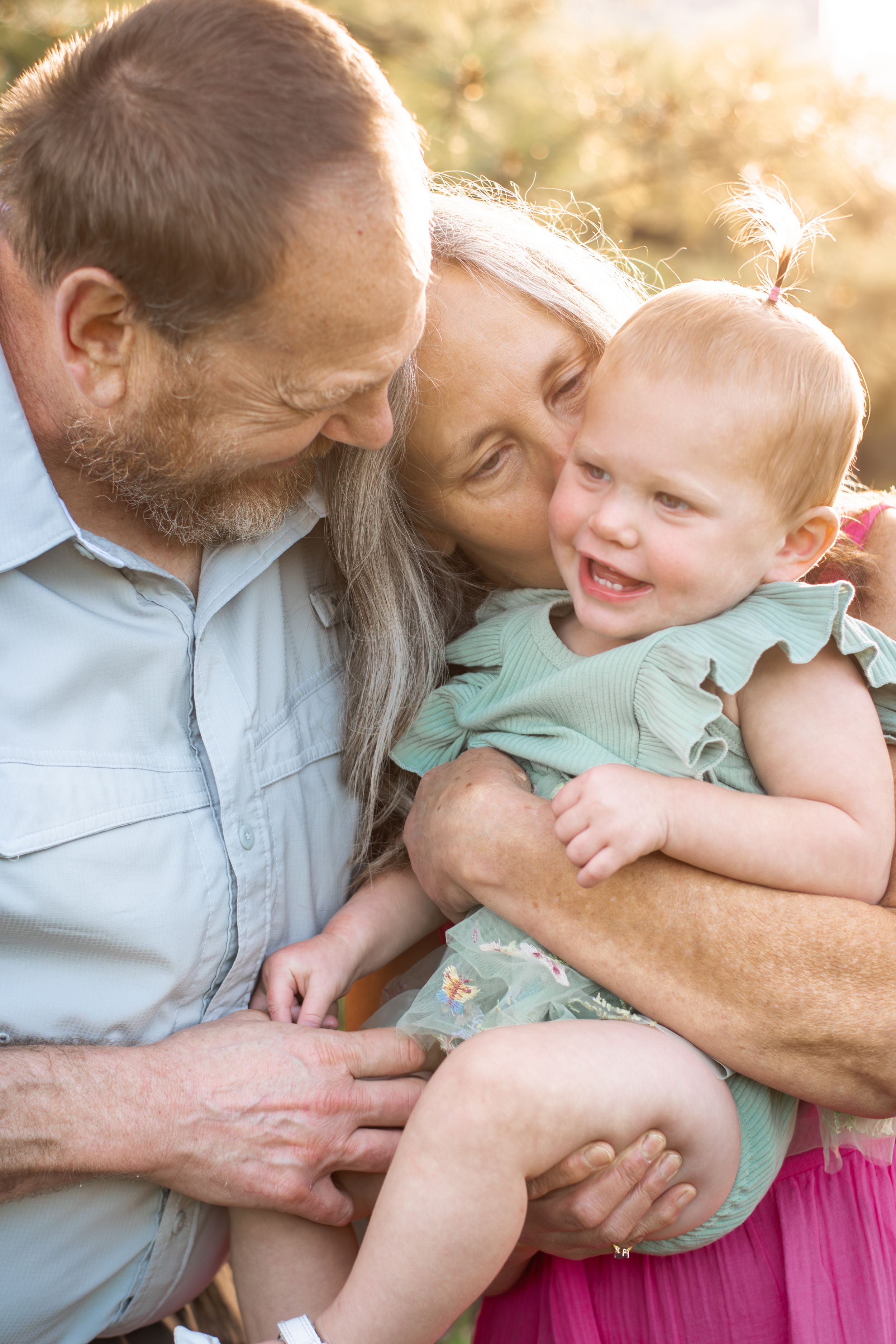 Grandparents cuddling and kissing a laughing red-haired baby girl outdoors in warm sunlight.
