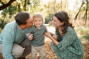 Smiling parents kneeling to kiss their toddler son on cheeks in an autumn park.