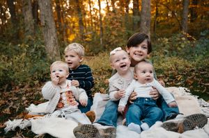 Five young children sitting together on a blanket surrounded by colorful autumn foliage.