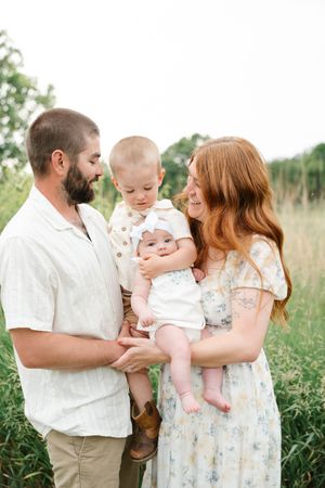 Smiling family of four with toddler and newborn posing together in lush green meadow.