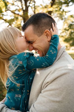 Adorable young blonde girl kissing her laughing father on the nose in a sunny park.