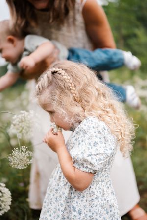 Curly blonde toddler girl in floral dress smelling wildflowers in a meadow with mother and baby in background.