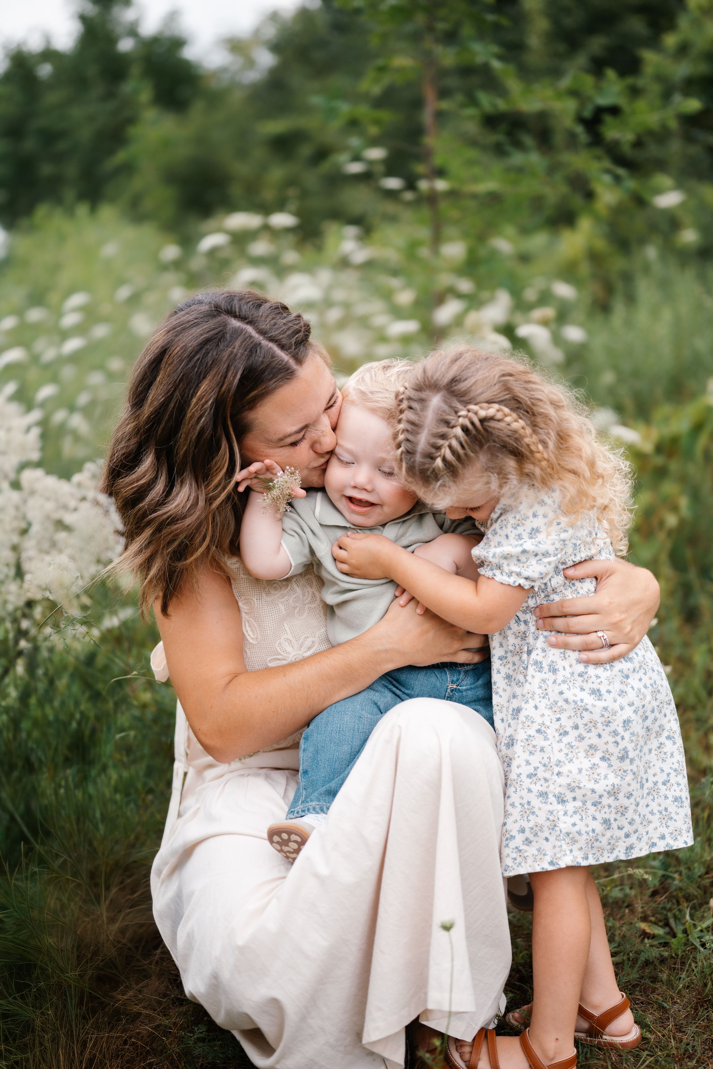 A mother kisses her laughing toddler boy while her young daughter hugs him in a wildflower field.