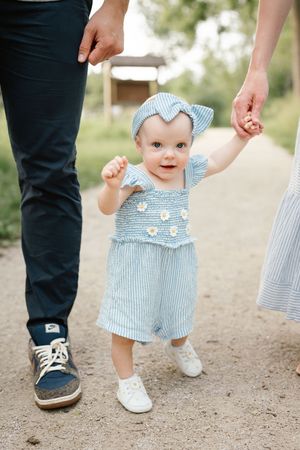 Adorable baby girl in blue floral dress taking first steps while holding parents hands outdoors.