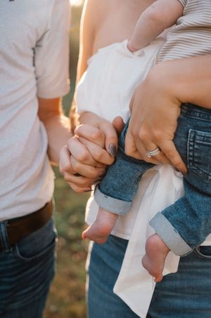 Parents holding hands while cradling a newborn baby outdoors in warm golden light.