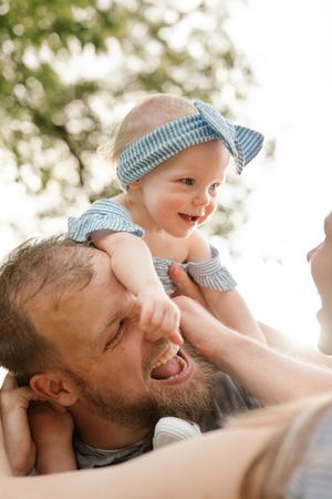 Joyful baby girl in blue outfit laughing while being lifted on fathers shoulders in park.