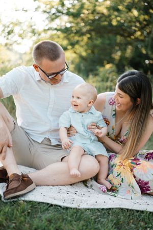 Family with baby sitting on blanket in park, parents smiling at laughing infant outdoors.