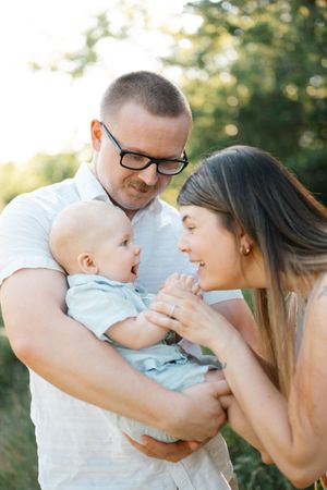 Parents holding baby outdoors, father in glasses smiling at infant while mother leans in close.