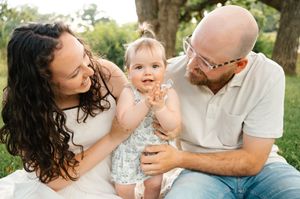 Loving family with baby girl sitting outdoors under green trees.