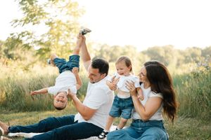 Happy family sitting outdoors in a sunny field, dad playfully holding toddler upside down while mom cradles baby.
