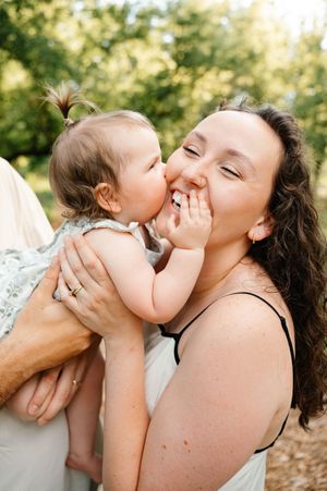 Baby girl giving sweet kiss to smiling curly-haired mother outdoors.