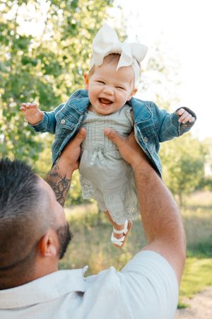 Joyful baby girl in denim jacket lifted by father in sunny green outdoor setting.