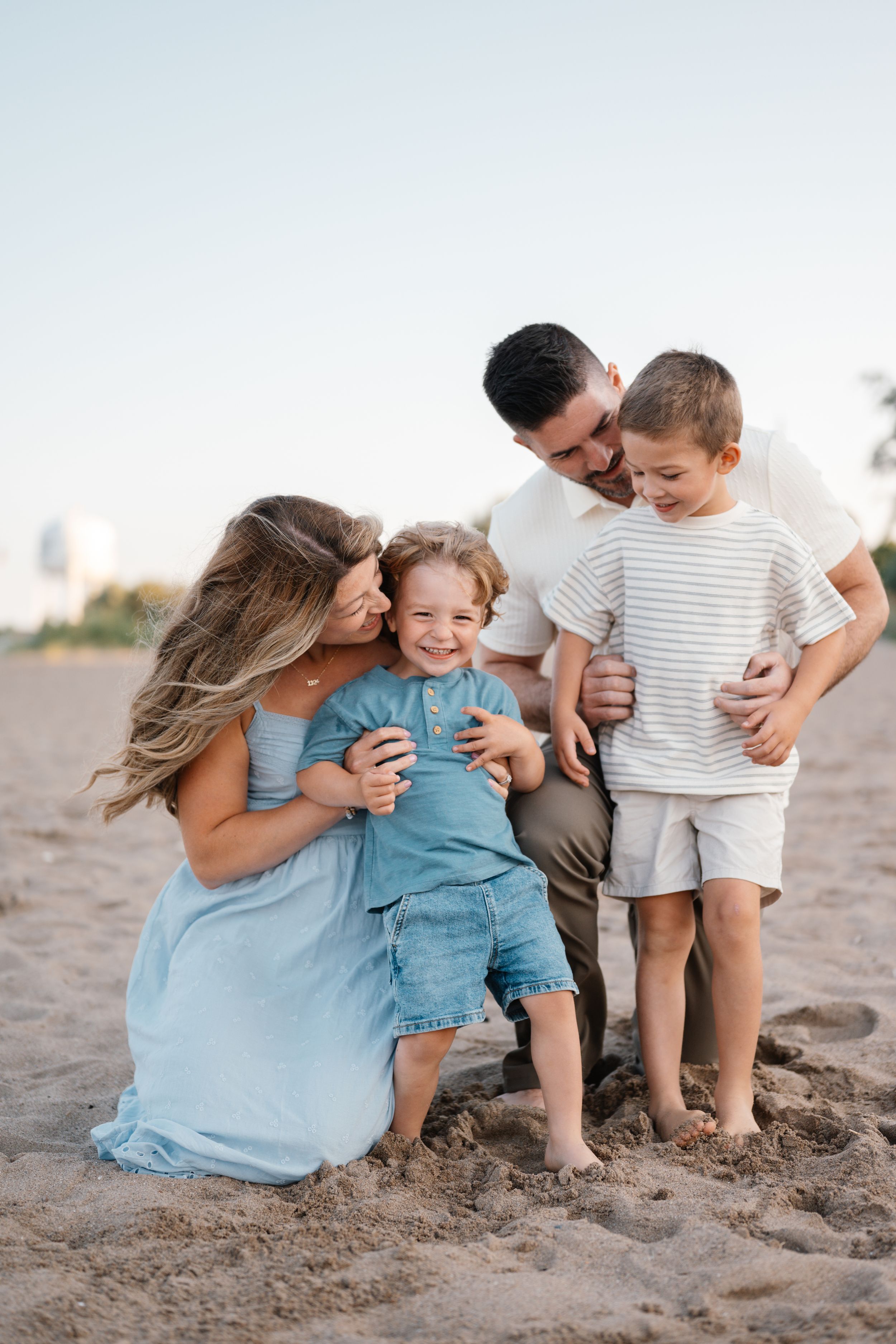 Happy family of four sharing joyful moments on a sandy beach, with parents hugging two young boys in casual summer outfits.