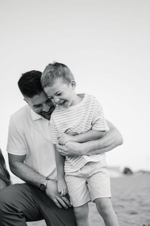 Black and white photo of a father hugging his laughing young son on a sandy beach.