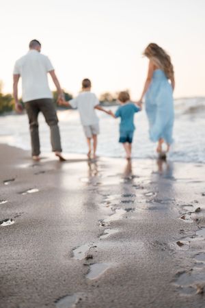 Family of four walking hand-in-hand along a sandy beach at sunset, leaving footprints in the wet sand.