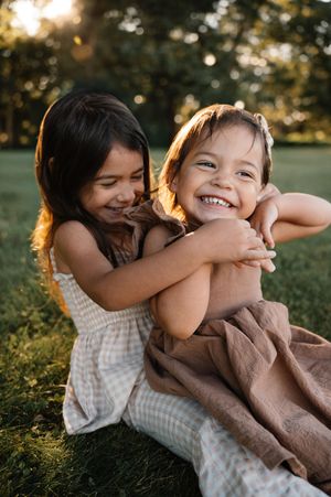 Two young sisters hugging and laughing joyfully together on green grass at sunset.