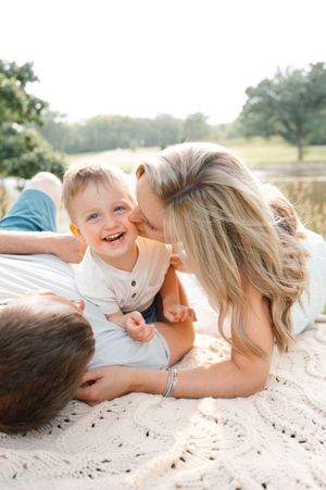 Happy toddler boy laughing while parents lie on blanket outdoors in golden sunlight.