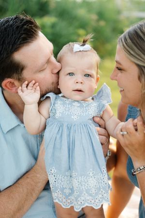 A couple lovingly holds their baby girl in a blue dress with a white bow, dad kissing her cheek outdoors.