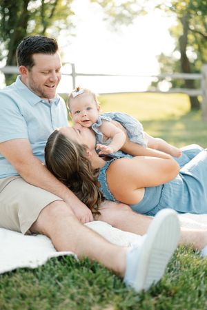 A happy family relaxes on grass outdoors, with parents smiling at their adorable baby girl in a blue romper.